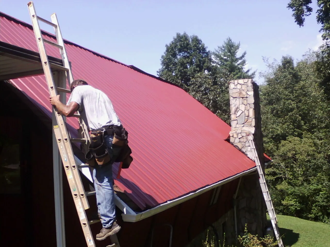 Roofer on a ladder beside a red metal roof and stone chimney — Soffit & Fascia Repair by Roofing Brothers in Stone Creek